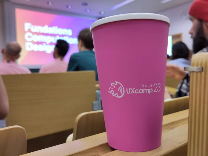Photo of cup with imprint 'UXcamp Europe 23'. Blurred background with conference context. 