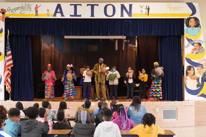 A Washington Performing Arts Teaching Artist ensemble is standing on stage at a school assembly. They are holding instruments and leading an interactive performance with student participants.