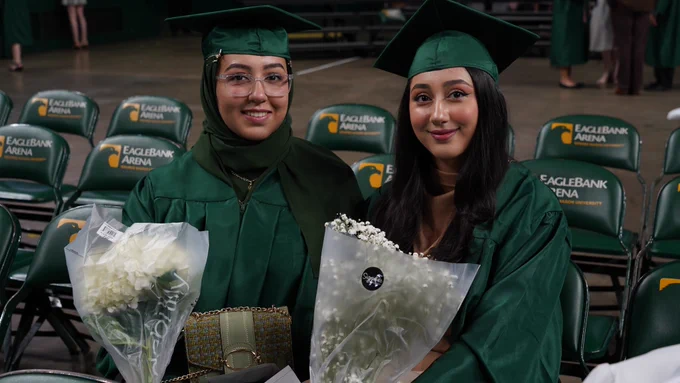 Two graduates in green graduation gowns show the flowers they received for the graduation ceremony.