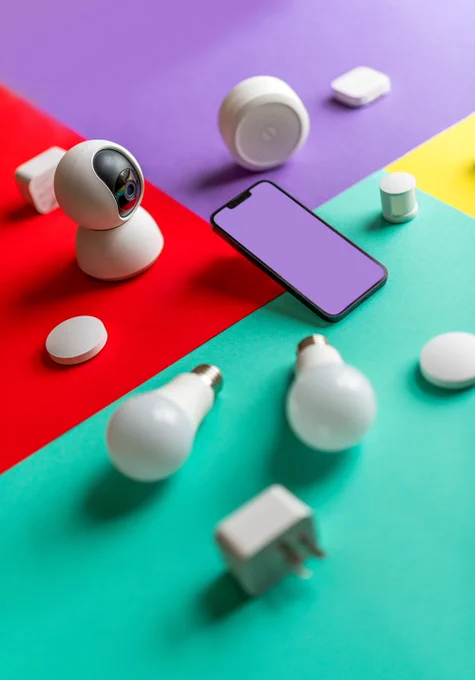 A table with various home automation technology displayed, including light bulbs, a speaker, motion sensors, a camera, and a smartphone.
