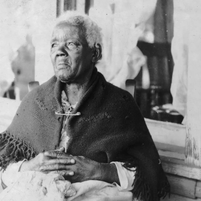 A portrait of an elderly Black woman, Sarah Gudger, who was reportedly 121 years old at the time the photo was taken to accompany the narrative she shared with the Federal Writers' Project. Her hair is short and white, her hands are in her lap with her fingers laced together and she is looking into the distance, to the left of the camera (her right). Her tattered shawl is safety-pinned together under her neck.