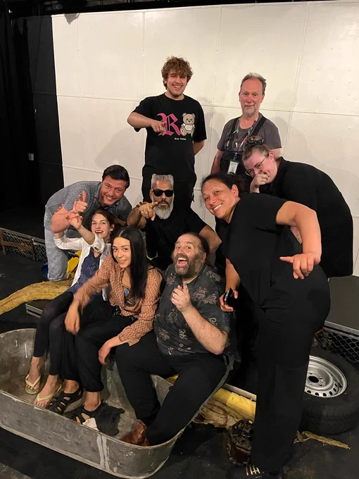 A group of nine diverse people sit and stand on an indoor stage. They smile and point at the camera. Three of them sit with their feet in a tin bath.