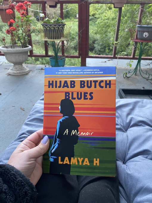 A hand holds the book Hijab Butch Blues with a stylized Hijabi turned away from the reader. The cover fades from oranges and reds so green. Author Lamya H writes her memoir under a pseudonym. Beyond the book the reader sits on a deck chair on a grey deck with red raining and green trees beyond. In front of and atop the railing are pots with plants and flowers