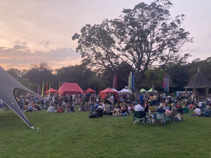 Grassy area with lots of people sat on deckchairs and on the grass, and standing up, with colourful gazebos and trees in the background, and with the sun starting to set in the sky.