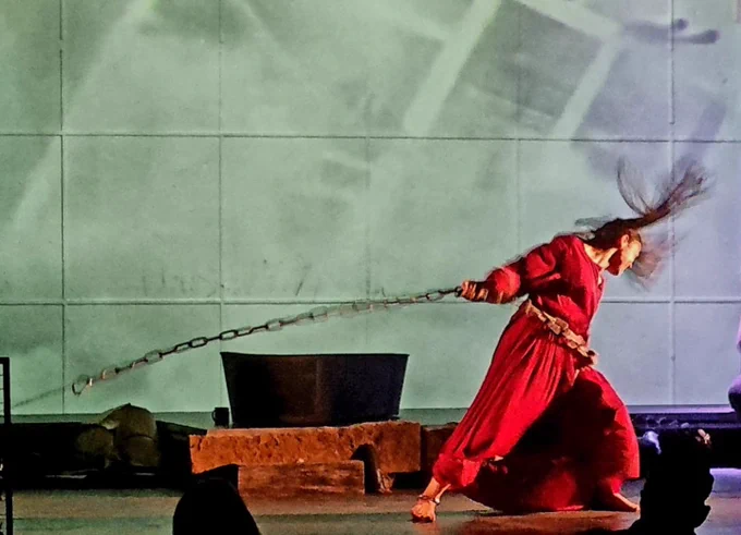 An indoor stage. A performer wearing a red dress pulls hard on a large chain to the right. Their long brown hair flies up. They have bare feet. Behind them to the left is a tin bath. On the back wall is a projected image of a falling building.