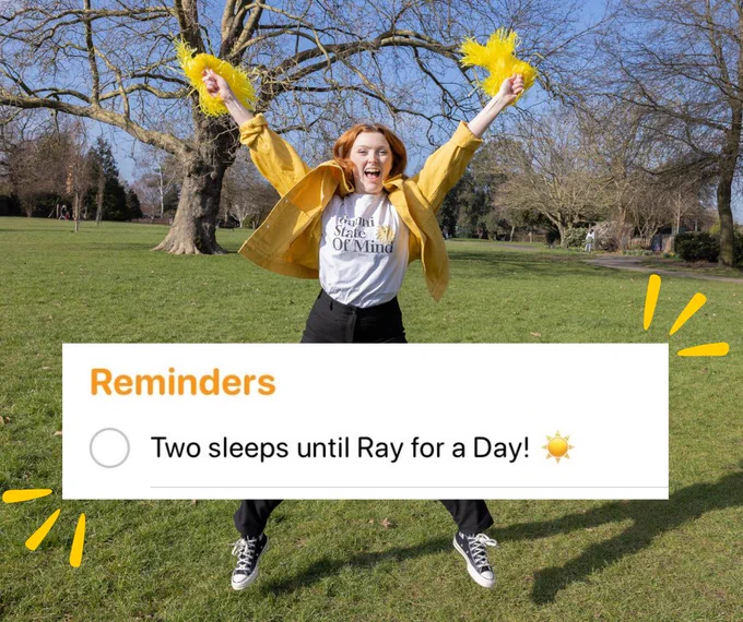 A red-haired girl does a star jump in a park. She is wearing a yellow jacket and is waving yellow pompoms. A graphic on the image reads: Reminders: Two sleeps until Ray for a Day.