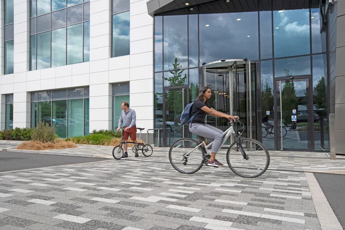 Person cycling past office building. Another stands in the background with a foldable bike.