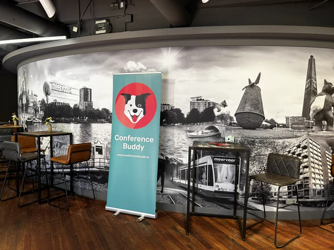 Picture of conference buddy Rollup and table standing at a half round wall with a black and white photo tapestry in the ground floor of the conference venue