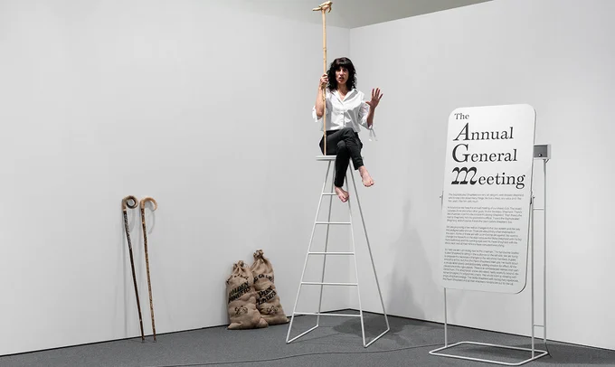 Image: Photo of the artist, barefoot, sitting on a very high stool facing us and holding what may be a walking-stick topped by what may be a little, prone, wooden human shape; the artist is apparently addressing an audience; leaning against a wall behind are two very long walking-sticks and two burlap sacks full of what may be apples • "New live artwork from acclaimed Irish artist Orla Barry blending autofiction and oral history to create a humorous, passionate monologue about the intersection of man and animal.  Wednesday, 7 June: 8.30pm  Thursday 8, June: 6.00pm  Friday, 9 June: 6.00pm  Saturday 10 June: 3.00pm  TO BOOK CLICK HERE    SPIN SPIN SCHEHERAZADE is a live artwork by Orla Barry, performed by Einat Tuchman her longstanding collaborator. It blends autofiction and oral history to create a humorous and passionate monologue. The performance is rooted in an artistic form of ‘barefoot anthropology’, recounting Barry’s tenuous interactions with the land, farming, man, and animal…"