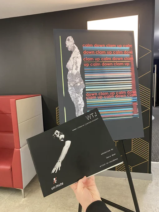 A hand holds up exhibition programme for Laura Dean’s exhibition un:mute, resident artist involved in the Women/Theatre/Justice project, in front of a piece of her work.