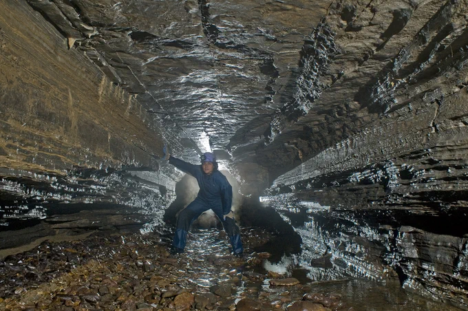 A woman in a jumpsuit and hard hat standing in the opening of a small cave. 