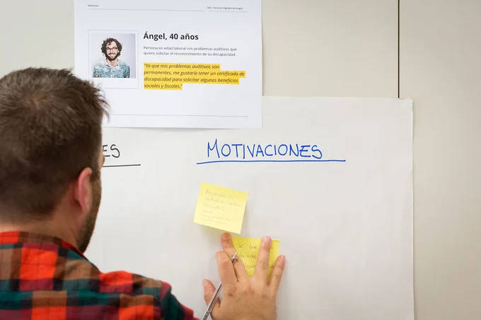 Un hombre de espaldas pega un post-it sobre un panel en blanco en el que se lee la palavra "Motivaciones"