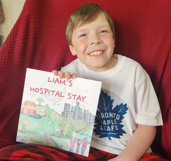 A young person named Liam, wearing a white Toronto Maple Leafs T-shirt, sits in a chair draped in a red blanket, holding a book he wrote called, Liam's Hospital Stay.