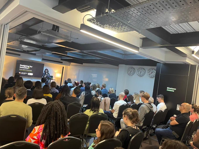 A large room filled with people sitting listening to a speaker at the front. 