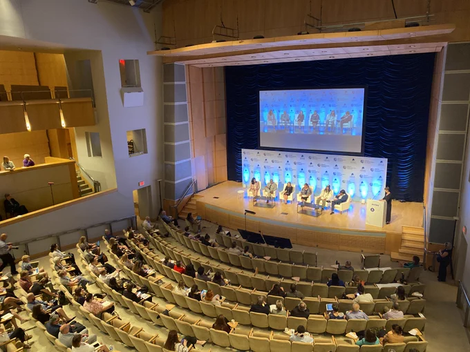 Six seated panelists on a stage in front of a projection address a seated audience.