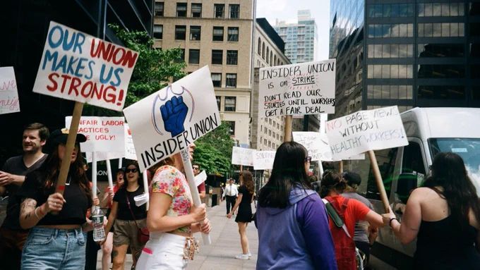 A group photo of people walking with picket signs on a sunny day in the city. On the far right a person holds a sign reading "OUR UNION MAKES US STRONG!" in all caps, the center person holds a sign with a blue fist inflame and reads "INSIDER UNION" in all caps, in the far right two signs are being held that read "INSIDER UNION is on STRIKE. DON'T READ OUR STORIES UNTIL WORKERS GET A FAIR DEAL." and "NO PAGE VIEWS WITHOUT HEALTHY WORKERS" 