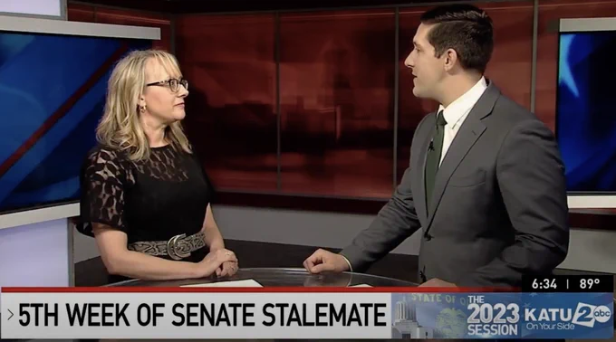 Photo of woman with blonde hair and glasses standing across from news anchor discussing local politics.