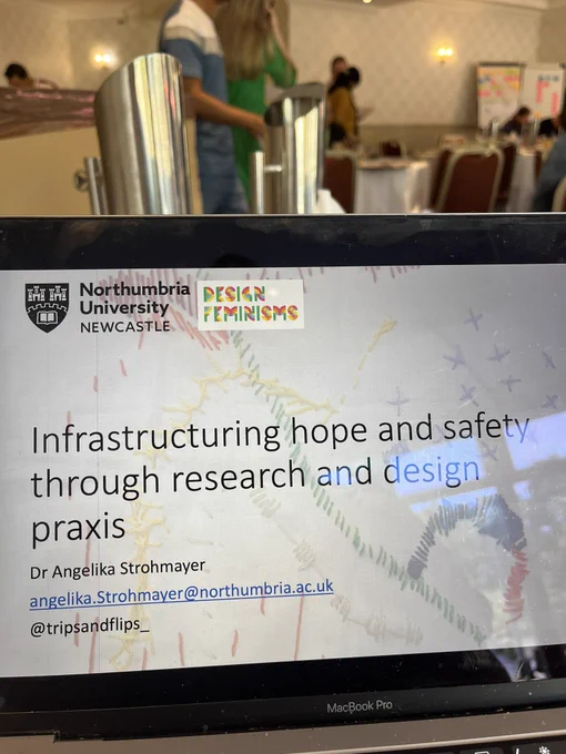 Photo of a laptop screen with a slide at full screen. The slide has a picture of white fabric with red, yellow, green, and blue embroidery mark-making stitches on it. The top left corner has the Northumbria University and Design Feminisms Research Group Logos. The title of the talk is: infrastructuring hope and safety through research and design praxis. The talk is by Dr Angelika Strohmayer with her email address and Twitter has file underneath her name