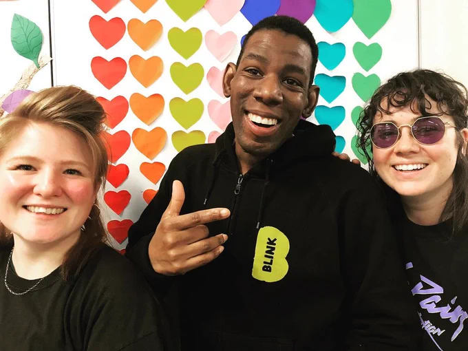 Image shows BLINK Facilitators Georgia (on the left, a white woman with ginger hair) and Kat (on the right, a white woman with dark, curly hair) and BLINK co-director Delson (centre, a black man with short, black hair, wearing a BLINK hoodie) smiling