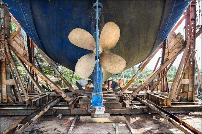 Image: Kevin O'Farrell: Repairing the keel on a 56’ wooden trawler, Hegarty’s Boatyard, 2014 – under what seems a huge hull we see two men working on a boat's keel; the large propellor and (possibly) rudder are closest to us; the hull is supported by a complex arrangement of girders short, thick beams  • "The last surviving traditional wooden boatyard in Ireland.This body of documentary photography, developed over 20 years, celebrates Ireland’s disappearing maritime heritage and the skills of traditional wooden boatbuilders focusing on three generations of the same family working in Hegarty’s Boatyard, Oldcourt, Skibbereen, Co Cork. Kevin O’Farrell’s practice draws on the traditions of photojournalism and magazine reportage. The exhibition comprises many photographs of the restoration of the AK Ilen, the last of Ireland’s traditional wooden, ocean-going sailing ships, as well as a new set of photographs of the restoration of the Saoirse, a 42 foot ketch on which Connor O’Brien…"