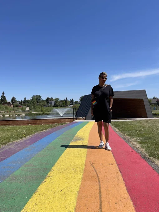 Aditi standing on a rainbow sidewalk with a fountain in the back. Aditi is wearing all black with a pride Fanny pack