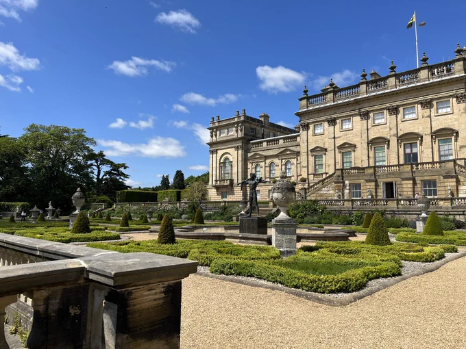 Harewood house viewed from the terrace, showing the beautiful gardens and the back of the house against a blue sky dusted with clouds.
