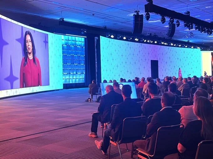 Picture of the stage at A23. Barbara Bouza, FAIA, on an LED screen and on the stage to the right