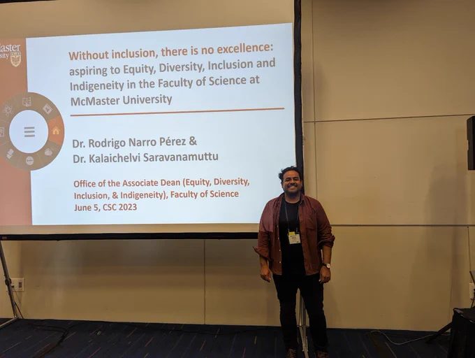 Rodrigo (wearing black pants and a black shirt with a burgundy shirt) standing next to a projector with a PowerPoint slide saying "Without Inclusion, there is no excellence: aspiring to Equity, Diversity, Inclusion and Indigeneity in the Faculty of Science at McMaster University by Dr. Rodrigo Narro Pérez and Dr. Kalaichelvi Saravanamuttu, office of the associate Dean (equity, diversity, inclusion and Indigeneity) 