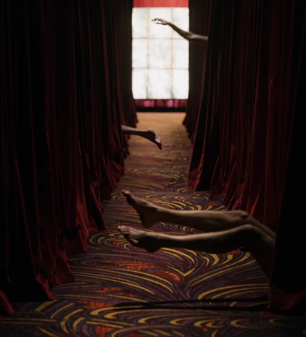 A dimly lit hallway walled by red velvet curtain, from which various limbs extend.