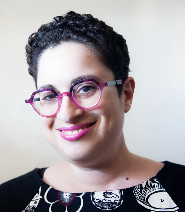 Anna Jacobson smiling - dark curly hair, brown eyes, pink lipstick, pink glasses and black shirt.