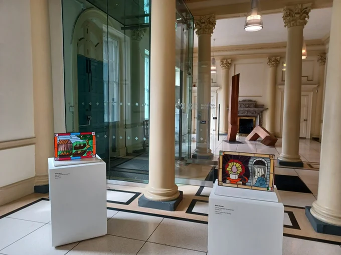 Image: photo of two stained-glass works, each about A2 size (?) on plinths in the entrance hall of the Hugh Lane; the closer work depicts a  lit candle in candle-holder on the left, and a rough-sleeper in a Georgian doorway on the right; the farther work suggests a rural landscape, or – more likely – an allotment, with plants growing in raised beds • "Solder: Global Connection through Art is the culmination of two years intense work by adult participants in the LYCS Global Citizenship project. The artists accepted the challenge to investigate and reflect upon Global Citizenship Education and the UN Sustainable Development Goals. Working with Helena Murphy in LYCS, there were many weeks of debate and reflection, after which the participants worked with artist Claire Halpin to bring their responses to life in stained glass.    At the outset of their project the artists visited the Hugh Lane Gallery where they were welcomed by Jessica O’Donnell, Head of Education and Community Outreach…"