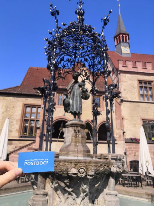 Göttingen's Gänseliesel (Goose girl) fountain in the main square in front of the old town hall. There is a blue card held in front reading "What would you change as a postdoc?" - more info about this much kissed statue is here: https://en.wikipedia.org/wiki/Gänseliesel. The address of this popular meeting place is: Markt, 37073 Göttingen