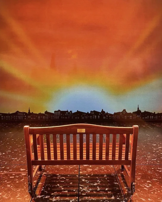 The back of an empty wooden park bench on stage. The bench faces a golden and orange sunrise over the rooftops. The plaque is at the top of the backrest in the centre. 