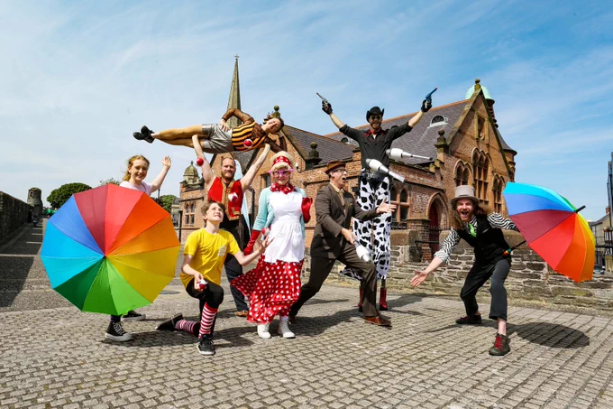 A group of eight circus performers gather on the City Walls outside the Old Cathedral School building. One of the performers is on stilts, two are juggling, one holds another performer above their head and two people are holding rainbow umbrellas.
