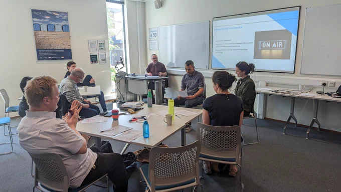 Inside a brightly lit room, seven of our researchers attend a session on engaging with the media. One of our researchers, sitting at the front, is rehearing live interview skills with one of the trainers. 