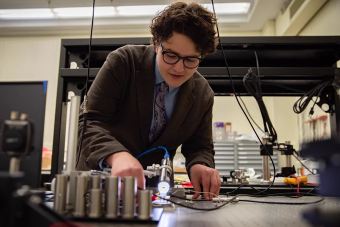 Man in a suit bends over a table with various extensions of wiring, a light source and other scientific instrumentation.