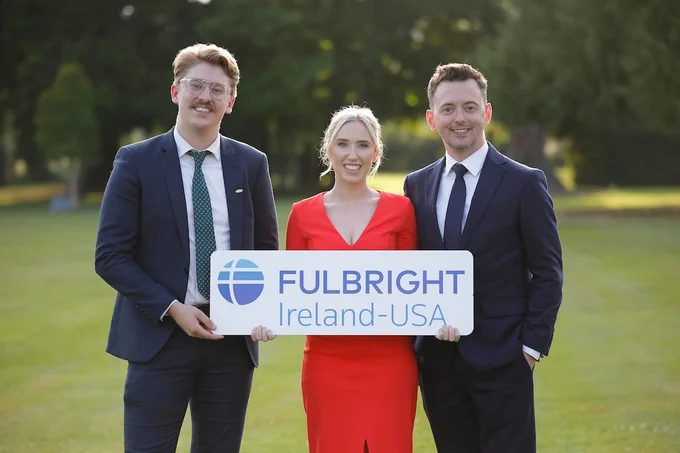 Photograph of the three award winners standing on a lawn outside holding a sign that reads FulBright Ireland-USA. All three are dressed formally.