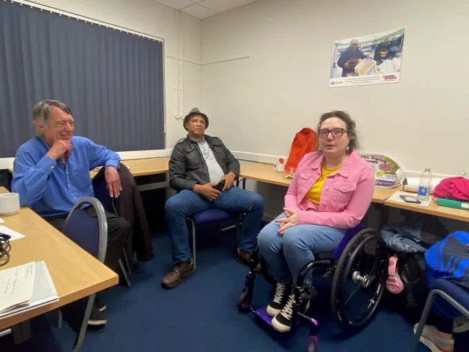 Three people sitting in a small room, all facing the camera. On the left is a man.  He is laughing.  In the middle is a second man.  He looks relaxed.  On the right is a woman, who has just noticed that a photo is being taken.  The men are sitting on chairs and the woman is sitting in her wheelchair.