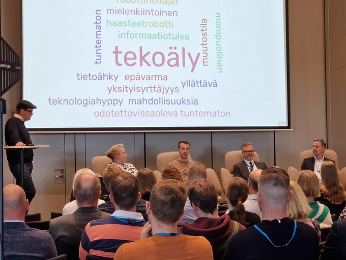 Paneelikeskustelijat ja fasilitaattori yleisön edessä. Taustalla sanapilvi, jossa yleisön kuvailemia sanoja tulevaisuudesta.