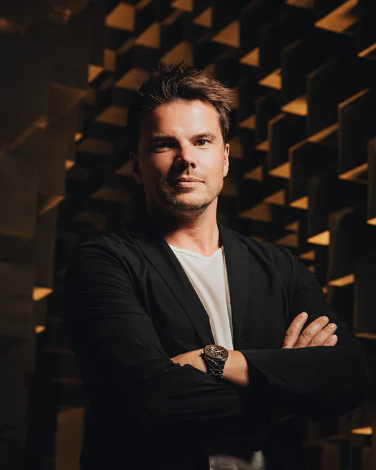 Danish architect bjarke ingels standing in front of an architecturally snazzy building wearing a dark suit and white t-shirt combo staring straight at the camera