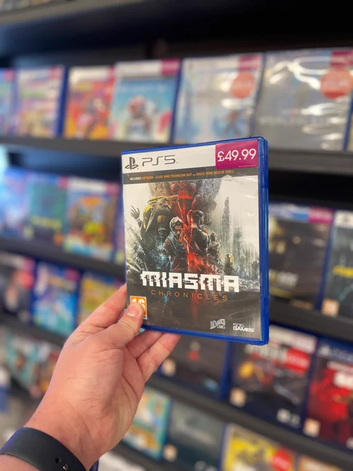 A hand is holding a box of a video game with the words Miasma Chronicles. There are other games blurred out in the background