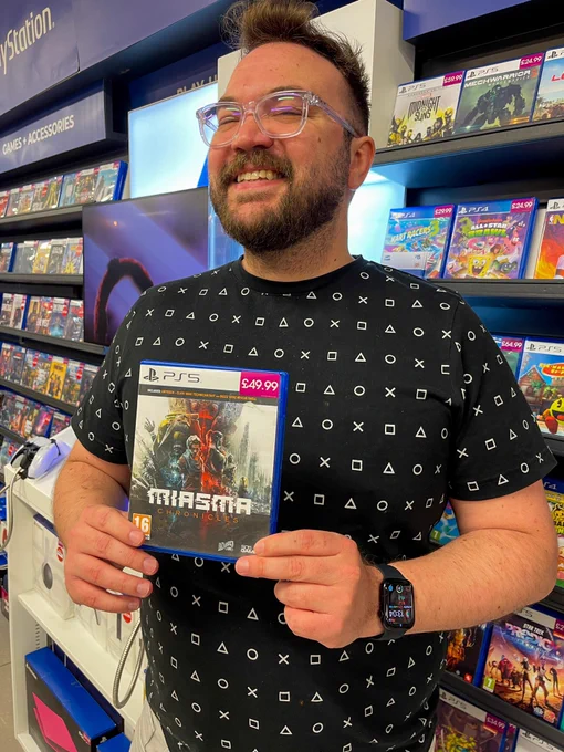 A man (me) is holding a video game box to his chest. On the box are the words “Miasma Chronicles”. The man has a black shirt with PlayStation symbols displayed and appears to be smiling.