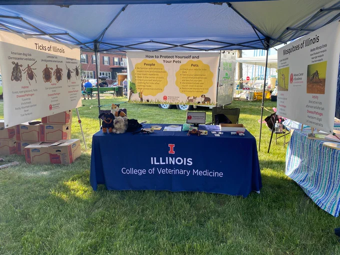 Tent with a table covered in a blue tablecloth that says "Illinois College of Veterinary Medicine" and hanging banners labeled "Ticks of Illinois", "Mosquitoes of Illinois", and "How to Protect Yourself and Your Pets"