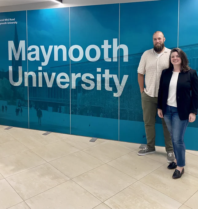 Lisa Carey and Cliff Thornton stand in front of a blue sign that says Maynooth University
