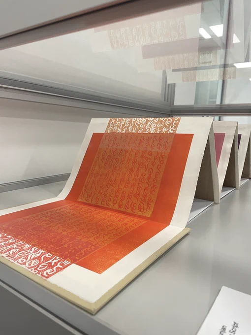 An image of an open accordion book within a glass case. The book shows a red page, with lino-print scripture printed onto the top in a block of orange, running the full width of the page.