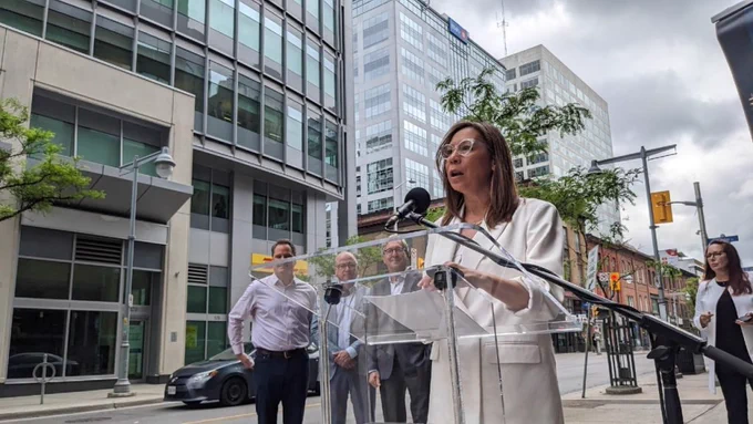 Sueling Ching speaks at a clear podium on the sidewalk outside a building downtown, with other high-rise buildings in the background