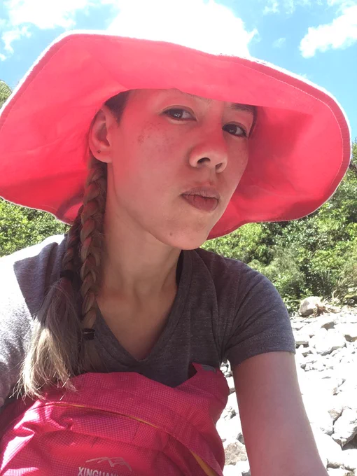 Selfie in a tshirt and a huuuge wide-brimmed hat that is white on top and bright pink underneath. Holding a bright pink backpack. Seated on a stony riverbank with green brush and blue sky.