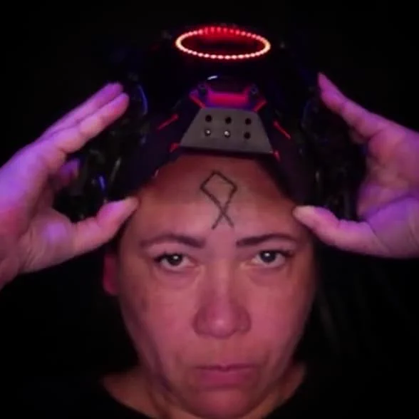 The face of a person with brown skin. They look intensely into the camera. On their forehead is a symbol drawn in black. It is a diamond with an upside down v connected below it. Above, a pair of brown hands in purple light hold up a black helmet with a circle of small red lights on the visor.