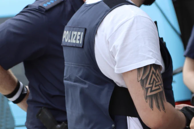 Ein Polizist im Schrägprofil von hinten, erkennbar am Schriftzug auf dem Rücken.
Am rechten Oberarm deutlich sichtbar ein Valknut