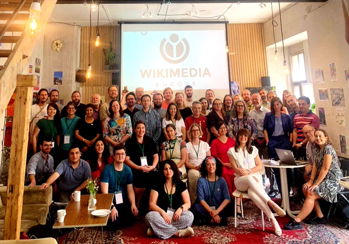 A group of about 50 häppy people from all over Europe in a group picture in front  of the Wikimedia Europe Logo